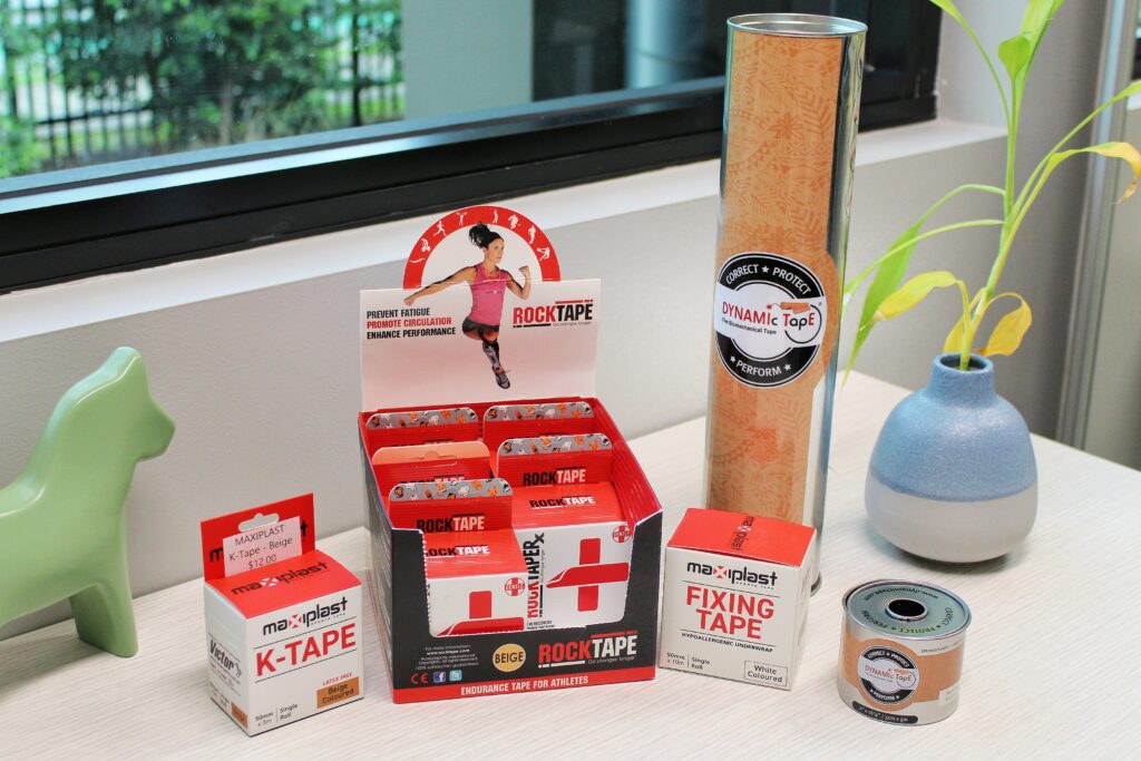 a display of various therapeutic tapes used in physiotherapy for hypermobility, including RockTape, Dynamic Tape, Maxiplast K-Tape, and Fixing Tape, arranged on a clinic desk